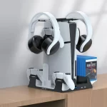 Green Lion PS5 cooling and charging station shown in a gaming setup on a wooden desk, holding a PS5 console, two controllers, two headsets, a media remote, and several game cases.
